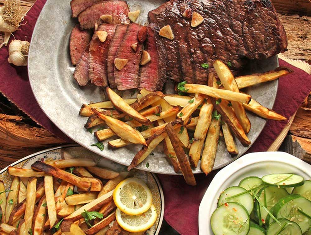 Grey plate with cut up steak and fries, surrounded by another grey plate with fries and two lemon slices and a white bowl with cut up cucumbers.